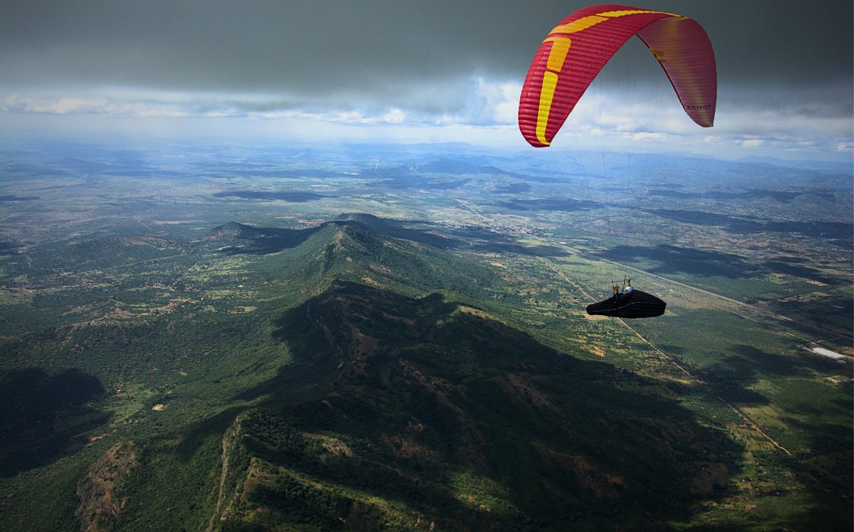 A red and yellow paraglider high above a steep green ridge with plains stretching to the horizon