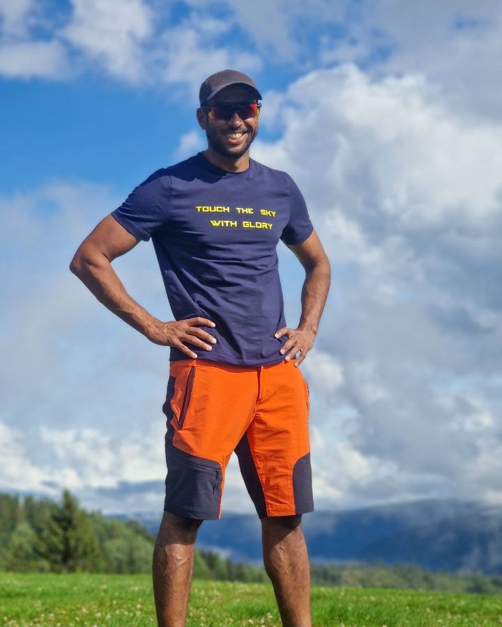 Aninder Singh standing on a green hillside in a Touch The Sky With Glory t-shirt