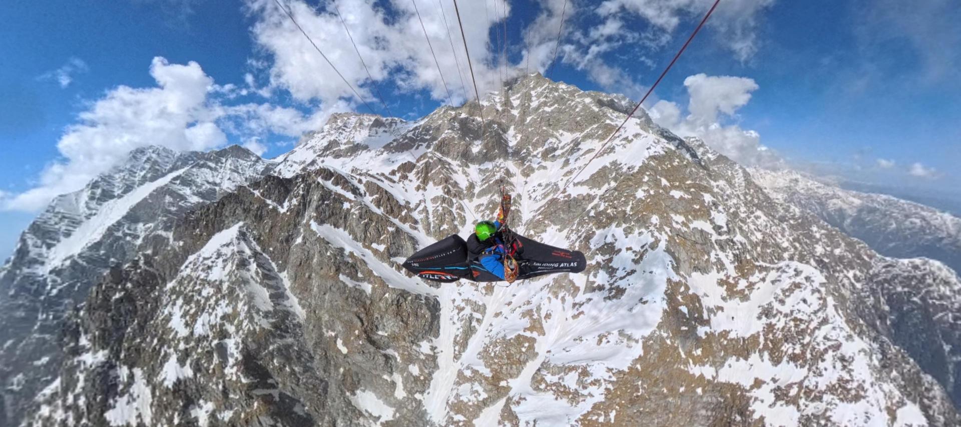 Aninder flying a paraglider above snow-covered peaks, the Paragliding Atlas name on his harness